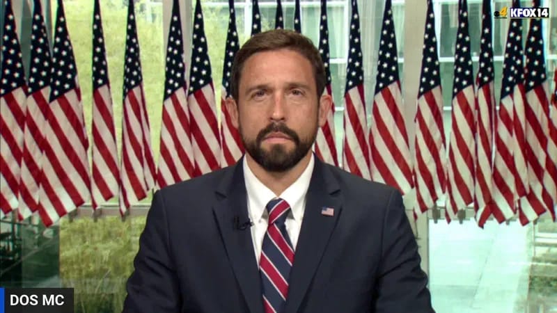 Man in suit with American flags behind him, likely discussing visa and immigration issues.