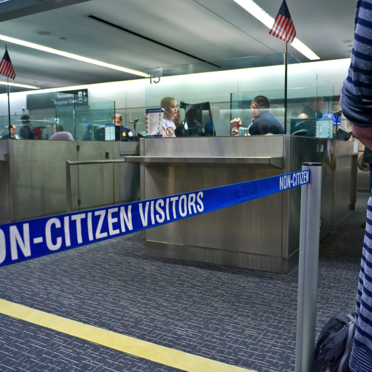 Non-citizen visitors line at airport immigration; Trump visa crackdown context.