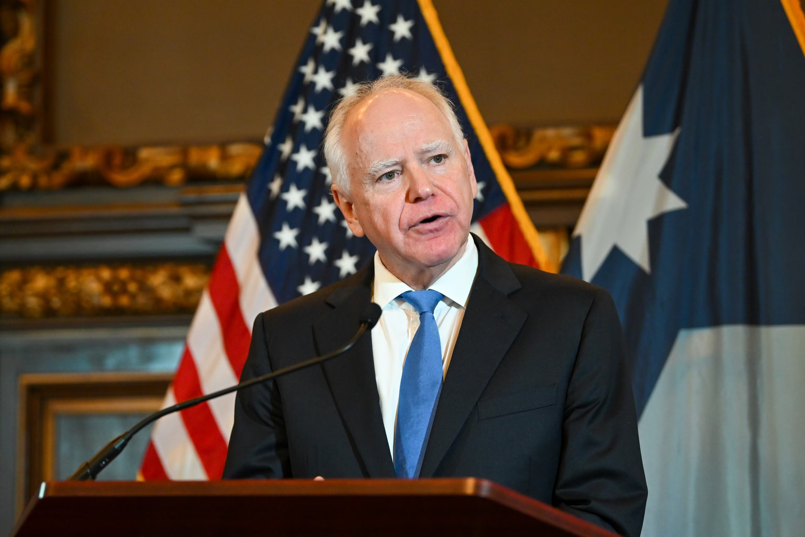 Minnesota Governor Tim Walz speaks at a podium with US and Texas flags behind him.