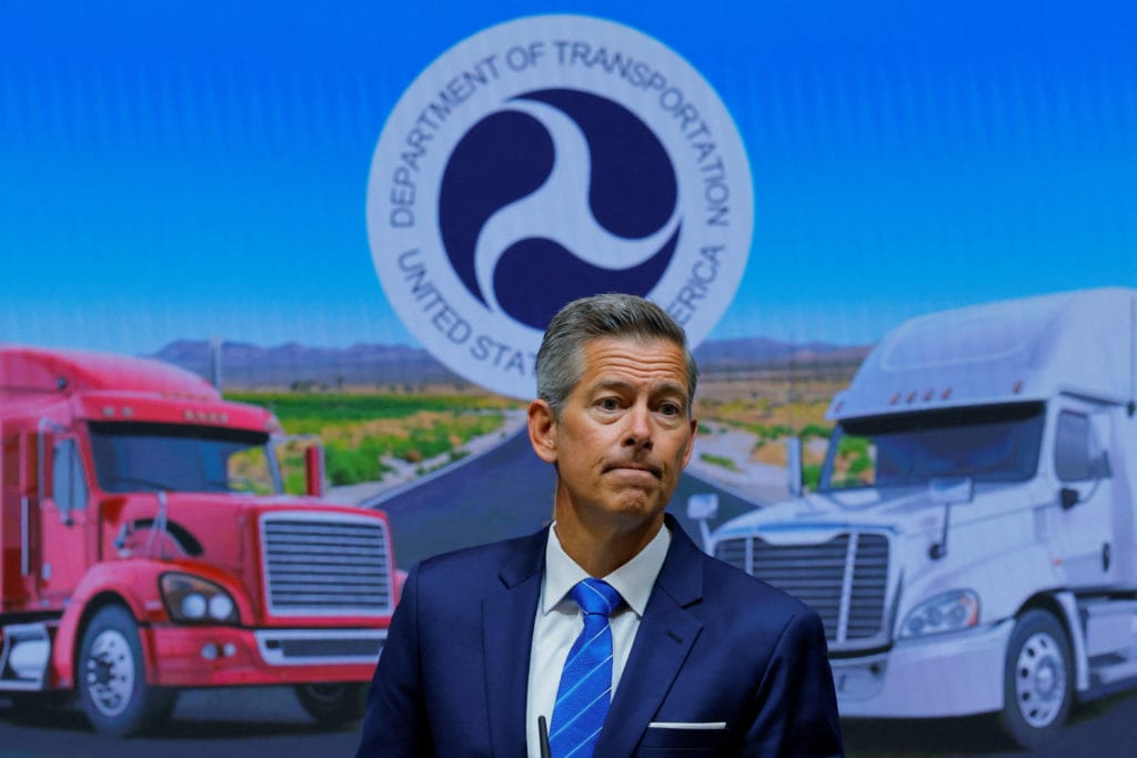 US Transportation Secretary Pete Buttigieg at a news conference with truck imagery.