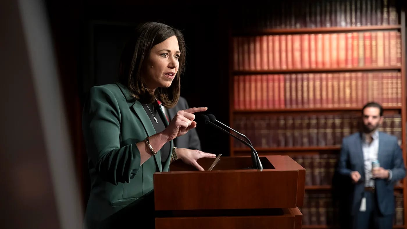 Senator Britt speaks at podium with bookshelves behind her